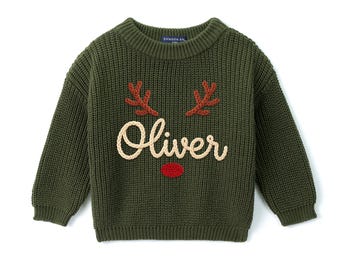 May include: An olive green long-sleeve knit sweater featuring a crew neckline. The front displays embroidered brown reindeer antlers, a red nose, and the name Oliver in cream-colored script lettering. The garment has a ribbed texture.