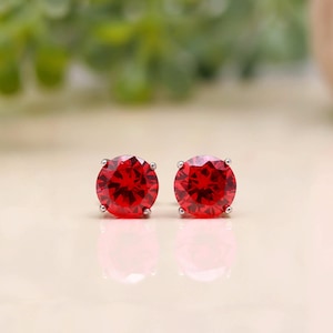Puede incluir: Un par de pendientes de botón con piedras preciosas rojas de corte redondo engastadas en una montura de metal plateado de cuatro garras. Los pendientes están colocados uno al lado del otro, mostrando una superficie facetada que refleja la luz.