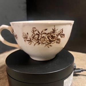 May include: Cream-colored teacup with a brown floral pattern. The cup has a curved handle and a slightly distressed appearance. The floral design includes roses and other flowers.