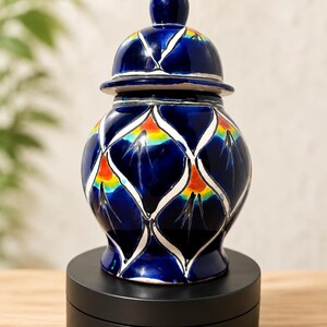 May include: A glossy ceramic jar with a removable lid, featuring a deep blue base color. The surface is decorated with a repeating white lattice pattern, with each section containing a vibrant orange and yellow gradient accent and black lines.