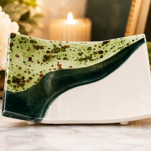 May include: A curved ceramic napkin holder featuring a glossy finish with a white base, a dark forest green wave-like accent, and a light sage green upper section speckled with small brown dots, supported by small feet on the bottom edge.