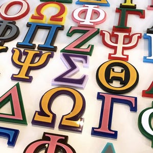 May include: A selection of brightly coloured Greek letter symbols, including red, blue, yellow, green, and pink. The letters are arranged on a white surface, each with a distinct outline and colour combination. Suitable for sorority or fraternity decoration.