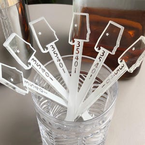 May include: Clear acrylic drink stirrers with an Alabama state outline and a star, each with the number "35401". The stirrers are in a clear glass with a silver cocktail shaker and a brown liquid in the background.