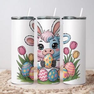 May include: White tumbler with a cartoon cow wearing bunny ears, holding a decorated Easter egg. The design includes colorful Easter eggs, tulips, and green grass. The tumbler has a silver lid and a white straw.