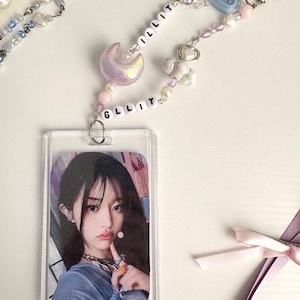 May include: A clear plastic ID holder with a photo of a person, attached to a beaded lanyard. The lanyard features white letter beads spelling "GLLIT" and "ILLIT", along with iridescent moon and heart charms, and a silver clasp.