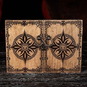 Puede incluir: Una caja de madera con un diseño decorativo, con dos puertas con bisagras ornamentadas y tiradores de anillo. La caja tiene un acabado marrón claro con un patrón grabado en marrón oscuro de una estrella y formas geométricas.