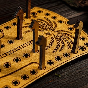 Può includere: Un gioco da tavolo in legno con un design decorativo a forma di grano e diversi pioli verticali. Il tabellone è marrone chiaro con dettagli marrone scuro, inclusi i pioli e il bordo. Il gioco è ambientato su una superficie di legno scuro.