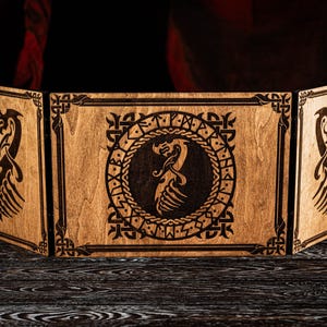 May include: A wooden tabletop game screen with a tri-fold design. The screen features intricate laser-cut dragon and rune designs. The wood has a natural finish, and the screen has metal ring handles.
