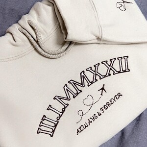 Può includere: Una felpa con cappuccio beige con un design ricamato marrone che dice "II.MMXXII" con un piccolo aereo e un cuore sotto. Il testo "ALWAYS & FOREVER" è anche ricamato sotto il design.
