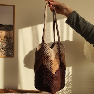 May include: A handmade crochet tote bag in shades of brown, beige, and tan. The bag features a chevron pattern and long handles. The bag is held up by a person's hand.