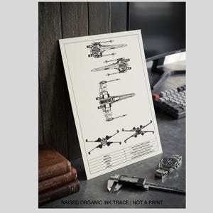 May include: A black and white schematic print of X-wing starfighters. The print is on a cream-colored paper and is leaning against a wooden surface. A watch, a caliper, and a stack of books are in the foreground. The text "RAISED ORGANIC INK TRACE | NOT A PRINT" is at the bottom.
