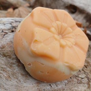 May include: A round, peach-colored soap bar with a flower design. The soap is sitting on a piece of wood.