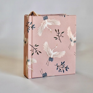 May include: A hardcover book with a dusty pink cover featuring a repeating pattern of white cranes in flight and dark blue leafy branches. The spine is visible on the left side, and a small decorative gold-toned metal accent sits at the top edge.