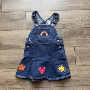 Puede incluir: Vestido peto de mezclilla azul con diseños bordados de arcoíris, flores, corazones y sol. Cuenta con tirantes ajustables, botones metálicos y falda plisada. Un atuendo colorido para un niño pequeño.