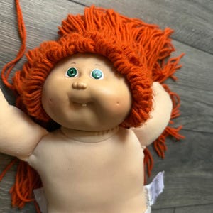May include: A vintage Cabbage Patch Kid doll with a soft, flesh-toned face, green eyes, and a tuft of orange yarn hair. The doll has a small, open mouth with a single tooth. The doll's body is made of a soft, beige fabric.