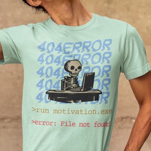 Op de afbeelding: Een lichtgroen T-shirt met een skelet dat aan een computer zit met de tekst "404ERROR" die meerdere keren wordt herhaald. De tekst onder het skelet luidt "run motivation.exe" en "error: File not found".