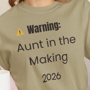 May include: A light olive green t-shirt with the text "Warning: Aunt in the Making 2026". The shirt has a yellow exclamation mark symbol above the word "Warning". The shirt is a crew neck style.