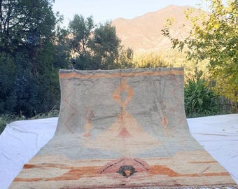 May include: A large, rectangular rug with a geometric design. The rug features a light gray background with orange, pink, and black geometric shapes. The rug has a fringe along the bottom edge and is displayed outdoors.