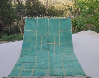 May include: A turquoise rug with a geometric design of beige lines and shapes. The rug has a fringe along the bottom edge. The rug is displayed outdoors, with trees and a mountain in the background.