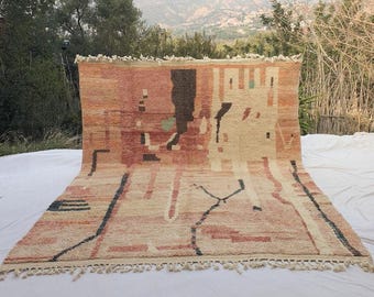 May include: A large, rectangular rug with a geometric design in shades of beige, red, and brown. The rug features black linear accents and a fringe along the bottom edge. The rug is displayed outdoors.