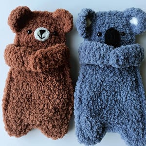 Puede incluir: Dos peluches hechos a mano: un oso marrón y un koala azul. El oso tiene una cara bordada en blanco y negro. El koala tiene una nariz negra y manchas blancas en las orejas. Ambos están hechos de un material suave y texturizado.