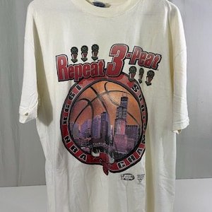 May include: Cream-colored t-shirt featuring a basketball design with the text "Repeat 3-Peat" in red. The graphic includes a basketball, the Chicago skyline, and the words "NBA Finals" and "Chicago Bulls".