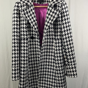 Könnte beinhalten: Ein schwarz-weißes Mantel mit Hahnentrittmuster und lila Futter. Der Mantel hat ein doppelreihiges Design mit zwei schwarzen Knöpfen und einem Reverskragen. Er hängt an einem Holzbügel.