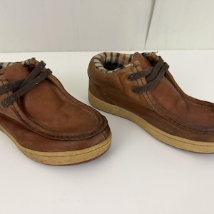 May include: A pair of brown leather ankle boots with a light brown sole. The boots have dark brown laces and a plaid fabric lining around the ankle. The shoes are designed for casual wear.