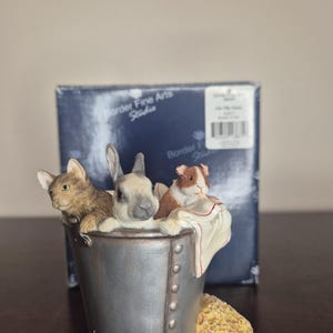 以下が含まれることがあります： 金属製のバケツに入った猫、ウサギ、モルモットを描いたBorder Fine Arts Studioの置物。動物たちは茶色とグレーです。白い布がバケツの縁にかけられています。