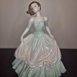 Puede incluir: Figura de porcelana de una mujer con un vestido fluido. El vestido es una combinación de blanco y verde pálido, con un escote corazón y guantes largos. La mujer tiene el pelo castaño y posa con un brazo extendido.