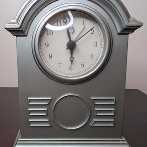 May include: A silver-colored, rectangular desk clock with a round clock face. The clock face is white with black numerals and hands. The clock has an arched top and decorative details on the front.