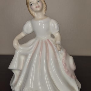 May include: A porcelain figurine of a young person in a white dress with pink accents. The figurine has blonde hair and is posed with one arm raised, holding the dress. The dress has a ruffled hem and pink ribbon detail.