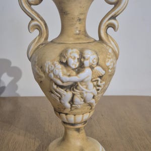 May include: A tan ceramic vase with two handles and a pedestal base. The vase features a relief of two cherubs holding flowers. The vase has a distressed finish and is suitable for decorative purposes.