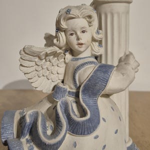 Puede incluir: Figura de ángel de cerámica con vestido blanco y detalles azules. El ángel tiene alas detalladas y sostiene una columna decorativa. La figura presenta una expresión serena y está adornada con detalles florales.
