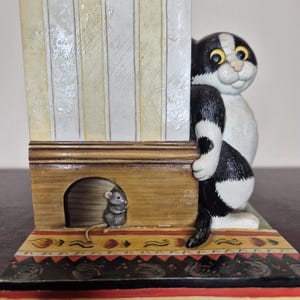 Peut inclure: Figurine fantaisiste représentant un chat noir et blanc regardant derrière un mur rayé. Une petite souris est dans une porte. La base présente un motif décoratif avec des accents rouges, noirs et jaunes. L'objet est décoratif.