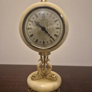 Op de afbeelding: Crèmekleurige quartz bureauklok met Romeinse cijfers en sierlijke details. De klok heeft een goudkleurige bekroning en een voetstuk dat wordt ondersteund door dolfijnvormige poten. De wijzerplaat heeft de tekst "Xavier of London" en "Quartz".