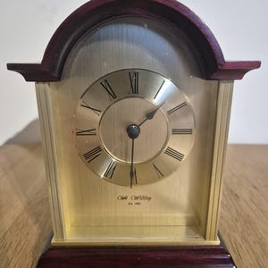 Peut inclure: Une horloge de cheminée rectangulaire avec un cadran doré et des chiffres romains. La partie supérieure et la base de l'horloge sont en bois foncé. Le cadran porte l'inscription "WR Widdop Est. 1883".