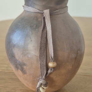 May include: A handcrafted, round, earthenware vase with a dark brown to tan gradient. A leather cord is tied around the neck, adorned with a wooden bead and decorative leather strips. The vase has a rustic, natural aesthetic.