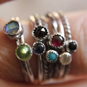 May include: A set of silver rings with colourful gemstones. The rings feature a variety of stones, including turquoise, green, red, black, and blue.