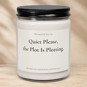 May include: A clear glass candle jar with a black lid containing white wax. The label reads: Bloomwick Soy Co, Quiet Please, the Plot Is Plotting, NATURAL SOY WAX BLEND | COTTON WICK. The candle is cylindrical with a simple, clean design.