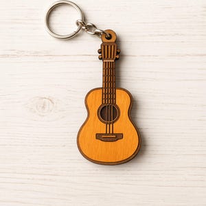 以下が含まれることがあります： 茶色のボディと薄い茶色の木目模様のついた木製ギターのキーホルダー。キーホルダーには、ギターの上部に銀色のキーリングが付いています。