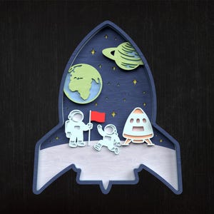 May include: A wooden artwork in the shape of a rocket. The design features astronauts, planets, and stars against a dark blue backdrop. The astronauts are holding a red flag, and a spaceship is also present.