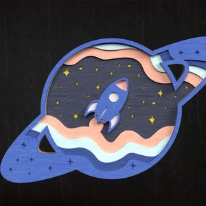 May include: A layered wooden artwork showing a blue rocket ship taking off into space. The piece features a dark blue background with stars, and a planet-like shape with layered colours of blue, peach, and white.