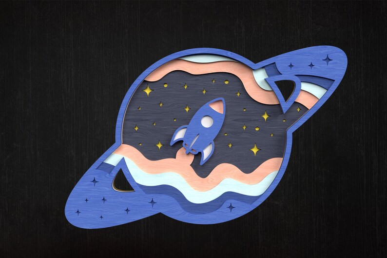 May include: A layered wooden artwork showing a blue rocket ship taking off into space. The piece features a dark blue background with stars, and a planet-like shape with layered colours of blue, peach, and white.