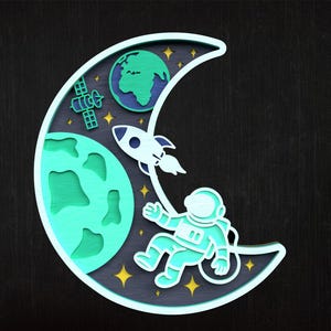 May include: A layered wooden artwork showing a space scene. A crescent moon shape frames an astronaut, rocket, Earth, and satellite. The artwork uses shades of blue, green, and yellow against a dark background.