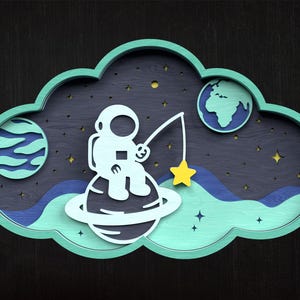 May include: A whimsical wooden art piece featuring an astronaut fishing for a yellow star from a planet. The design includes a blue and green cloud-shaped frame, planets, and stars against a dark background.