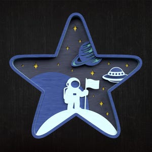 May include: A layered wooden star-shaped decoration with a space theme. It features an astronaut holding a flag on a planet, a planet with rings, and a UFO. The colour scheme includes shades of blue, white, and yellow.