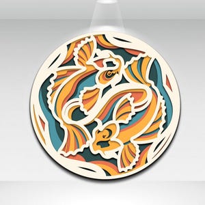 May include: A layered, circular art piece featuring two koi fish in shades of orange, yellow, and blue. The fish are set against a dark teal background, with a cream-colored outer frame. The design has a 3D effect, with each layer adding depth.