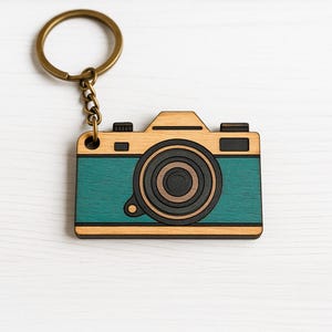 May include: A wooden camera keychain with a teal body and a black lens. The keychain is attached to a gold metal ring.