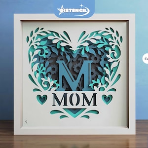 May include: A white frame with a blue and green heart-shaped design featuring the letter "M" and the word "MOM". The design is intricate and includes floral elements. The frame is 5 inches wide.